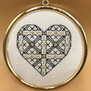 May include: A circular ornament with a gold frame, a white background and a black and gold embroidered heart design.