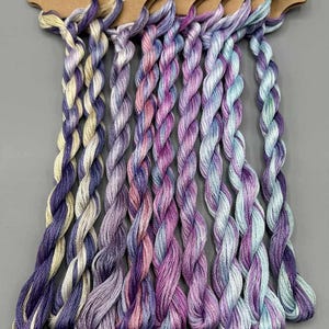 May include: A selection of hand-dyed embroidery floss in shades of purple, pink, and blue. Each skein is twisted and attached to a brown paper tag. The text "Light Fantastic: Set 2 of 4" is visible at the bottom.