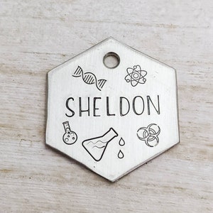 May include: A silver hexagonal dog tag with the name "SHELDON" engraved on it. The tag is decorated with science-themed symbols, including a DNA strand, an atom, a beaker, a test tube, and a biohazard symbol.
