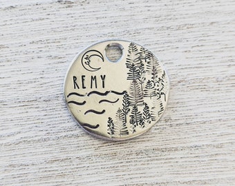 May include: Silver round pet tag with a crescent moon, waves, and a forest design. The tag is engraved with the name "REMY".