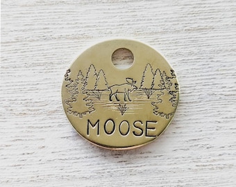 May include: A gold-colored metal tag with a moose silhouette and pine trees engraved on it. The tag reads "MOOSE" in all caps.