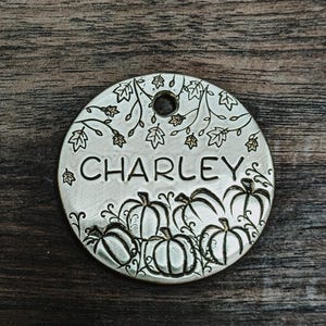 Peut inclure: Une médaille circulaire argentée pour animaux de compagnie avec le nom "CHARLEY" gravé. La médaille présente un motif de citrouilles et de feuilles, avec un trou en haut pour l'attache. Le fond est un grain de bois foncé.