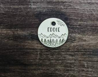 May include: Round, white metal dog tag with a mountain and tree design. The tag has the name "EDDIE" engraved on it.