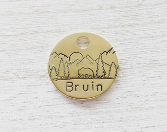 May include: A round, gold-colored pet tag with a mountain and forest scene etched into the surface. A bear walks across the landscape, with the name "Bruin" engraved below. The tag has a hole at the top for attachment.