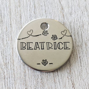 May include: A silver round tag with the name "BEATRICE" engraved in a decorative font. The tag is decorated with a bee and heart design.