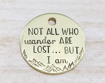 May include: A gold-colored round metal tag with the text "NOT ALL WHO wander ARE LOST... BUT I am" and a mountain and tree design.