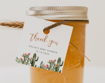 May include: A small glass jar with a silver lid, filled with a light orange liquid. A tag with the words "thank you" and "Helen's Baby Shower 10.18.2020" is tied to the jar with twine. The tag features a cactus design.