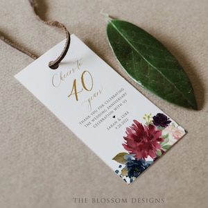 May include: A white tag with gold lettering that says "Cheers to 40 years". The tag is decorated with a floral design in shades of burgundy, pink, blue, and green. The text "Thank you for celebrating the wedding anniversary celebration with us Sarah & Luke 9.20.2022" is printed below the floral design. The bottom of the tag says "The Blossom Designs".