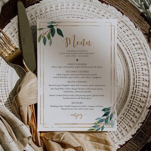 May include: A wedding menu card with a gold frame and watercolor greenery. The menu lists a multi-course meal, including lamb, chicken, and dessert. The card is placed on a decorative plate setting with a gold fork and knife.