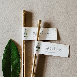 Könnte beinhalten: Zwei goldene Strohhalme mit befestigten weißen Papierfahnen. Die Fahnen haben grüne Aquarellblätter und goldenen Glitzer. Der Text auf den Fahnen lautet "Sip Sip Hooray" und "Linda's Bachelorette | 9.7.20".