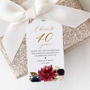 May include: A gift box wrapped in gold glitter paper, tied with a cream-colored satin ribbon. A white tag with gold script reads "Cheers to 40 years" and includes a floral design with burgundy and navy flowers.
