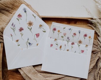 Puede incluir: Dos sobres blancos con un patrón floral en el interior. Los sobres están sobre una superficie de madera con una tela beige.