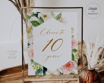 Könnte beinhalten: Ein gerahmtes Schild mit dem Text "Cheers to 10 years" in goldener Schrift, umgeben von Aquarell-Blumenmustern. Das Schild wird in einem goldenen Rahmen präsentiert, mit zwei Größenoptionen: 12,7cm x 17,8cm und 20,3cm x 25,4cm.