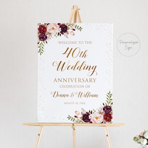 May include: A white sign on a wooden easel, with gold lettering announcing a 40th wedding anniversary celebration. The sign is adorned with floral arrangements in burgundy, pink, and white. The text includes the names Donna & William.