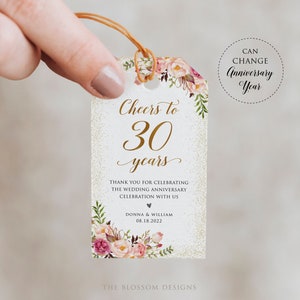 May include: A white tag with gold glitter and floral accents. The tag says "Cheers to 30 years" and "Thank you for celebrating the wedding anniversary celebration with us Donna & William 08.18.2022". The tag is tied with a brown string.