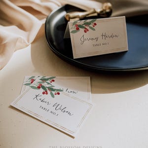 May include: Two white place cards with gold trim and a watercolor design of red berries and green leaves. The text on the cards reads "Jeremy Hardin, Table No. 1" and "Kabir Wilson, Table No. 1".