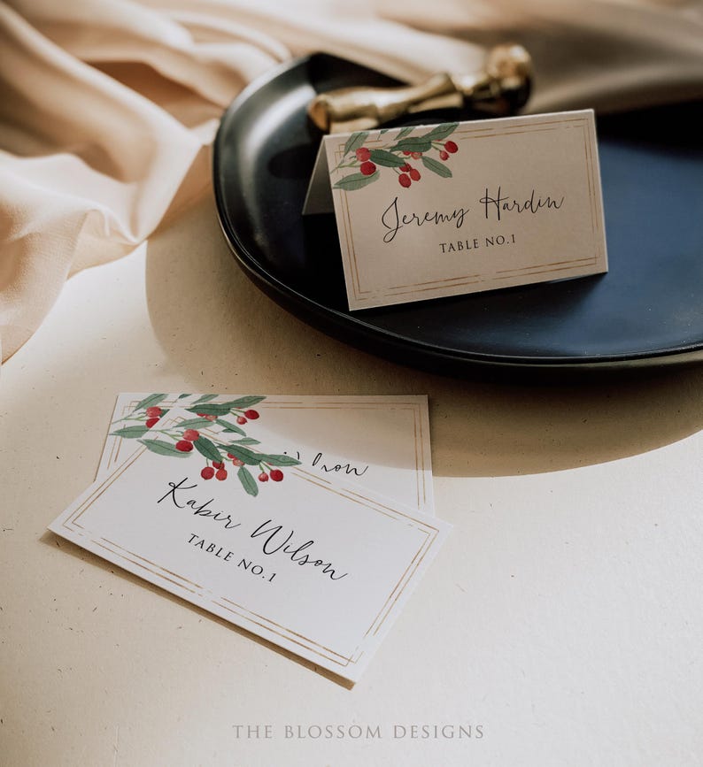 May include: Two white place cards with gold trim and a watercolor design of red berries and green leaves. The text on the cards reads "Jeremy Hardin, Table No. 1" and "Kabir Wilson, Table No. 1".