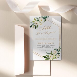 Puede incluir: Una tarjeta de invitaci&oacute;n blanca con detalles geom&eacute;tricos dorados y arreglos florales. El texto dice "Shhh... It's a Surprise!" y anuncia una celebraci&oacute;n del 50 aniversario para Karen & James. Incluye un sobre y una cinta.