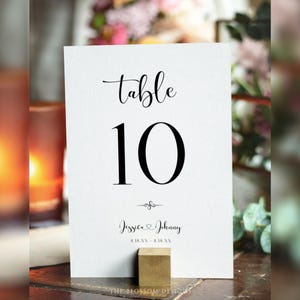 May include: A white table number card with the word "table" in elegant script above the number "10" in a bold font. Below, the names "Jessica & Johnny" are displayed, along with a date. The card is held by a small, gold-colored block.