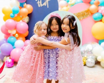 May include: Three young girls wearing pink, purple, and light pink dresses with butterfly designs stand in front of a colorful balloon backdrop. They are smiling and hugging each other.