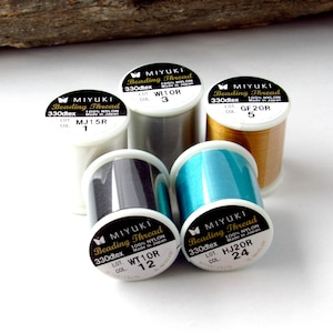 May include: Five spools of Miyuki Beading Thread made of 100% nylon, 330dtex. The spools are white, grey, gold, black, and teal. Each spool features a label with the brand name, material, and specific lot and color codes printed on the top.