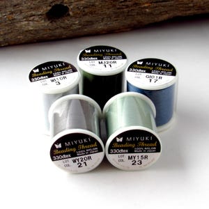 May include: Five spools of Miyuki beading thread made of 100% nylon. The spools are labeled with 330dtex and specific lot and color numbers including WI10R 3, MJ20R 11, GIO1R 17, WY20R 21, and MY15R 23. The threads are grey, dark green, blue, and light green.