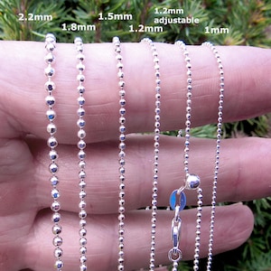 May include: Close-up of several silver ball chains of varying thicknesses, ranging from 1mm to 2.2mm. One chain has an adjustable clasp. The chains are displayed against a blurred green background.