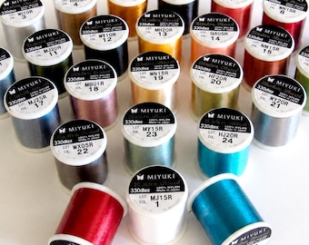 Puede incluir: Una colección de veinticuatro carretes cilíndricos de hilo de nailon Miyuki en una variedad de colores vibrantes. Cada carrete presenta una etiqueta blanca en la parte superior con la marca, 330dtex, 100% nailon, lote y código de color en negro.