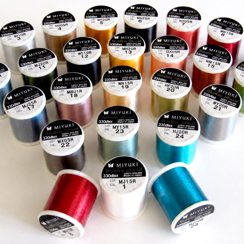 May include: A collection of twenty-four cylindrical spools of Miyuki nylon thread in a variety of vibrant colors. Each spool features a white label on top with the brand name, 330dtex, 100% nylon, lot number, and color code printed in black text.
