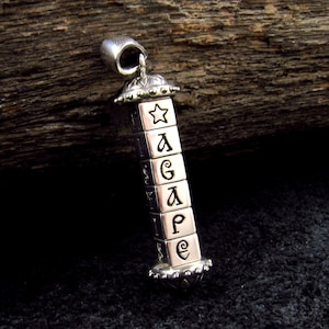 Puede incluir: Colgante de plata con la palabra "AGAPE" escrita en una serie de pequeños bloques cuadrados. El colgante tiene una parte superior e inferior decorativas, y una estrella es visible en el bloque superior.