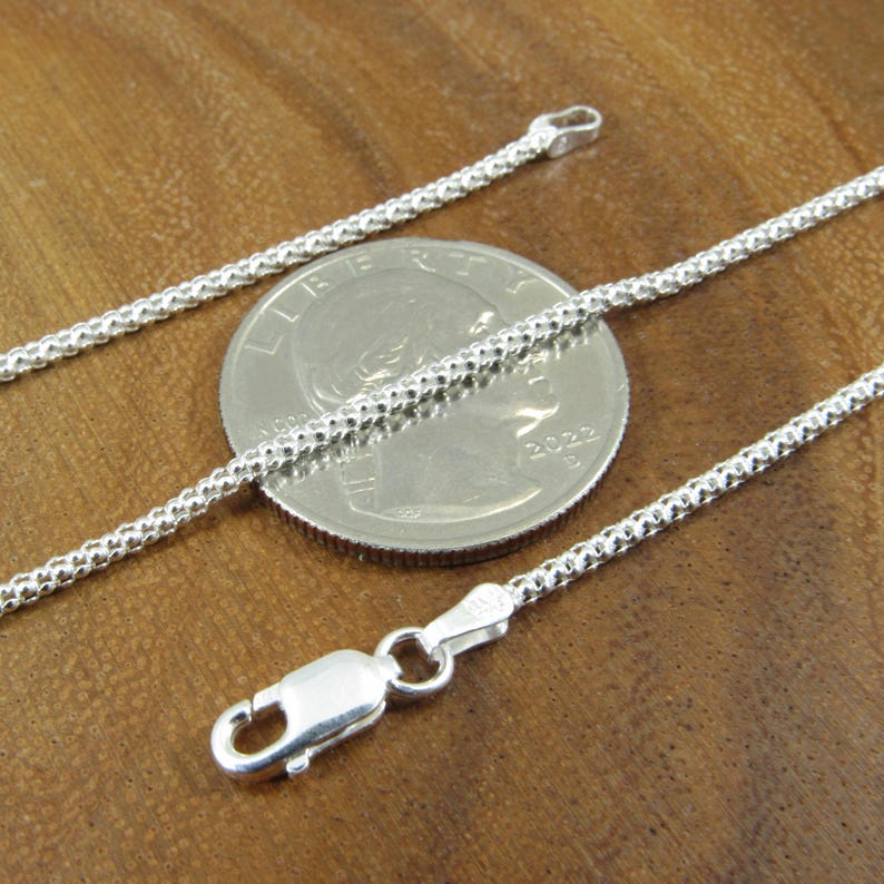 Op de afbeelding: Een zilveren ketting met een karabijn sluiting. De ketting wordt getoond naast een Nederlandse 1 euro munt voor een vergelijking van de grootte.