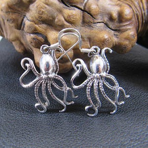 May include: A pair of silver-colored octopus earrings. Each earring features a detailed octopus design with multiple tentacles and a textured body. The earrings have a hook closure.