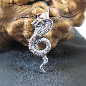 Puede incluir: Pendiente de cobra de plata con un diseño detallado y texturizado. El pendiente está en posición elevada con la cabeza girada hacia un lado. El pendiente está sobre una superficie de madera.