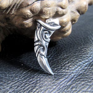 May include: A silver pendant shaped like a tooth with intricate, swirling designs. The pendant is made of metal and has a shiny finish.