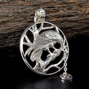 May include: A silver pendant featuring a detailed raven perched on a tree branch within a circular frame. The raven is depicted with intricate feather detailing. A small charm hangs from the bottom of the pendant. The background is a dark wood.