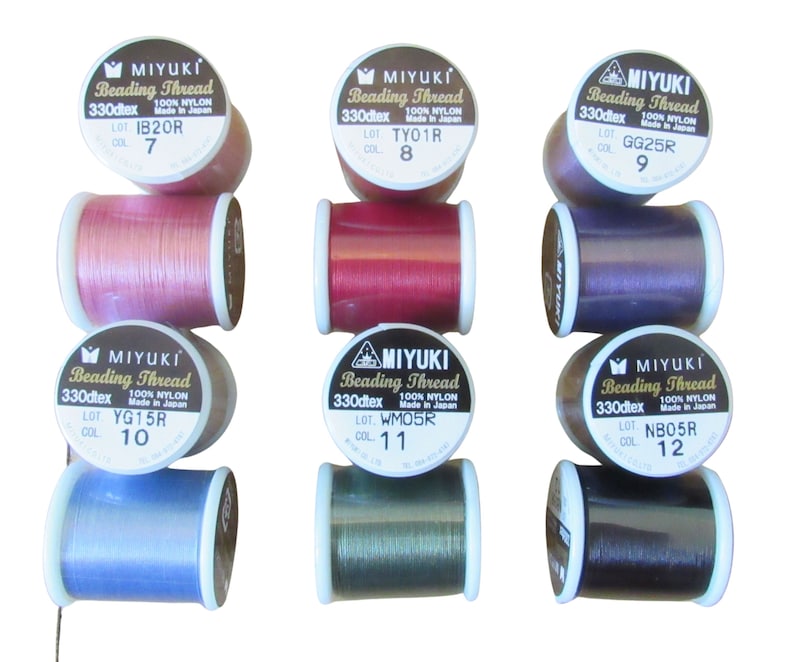 May include: Six spools of Miyuki Beading Thread arranged in two rows. Each spool features a white label with black text including the brand name, 330dtex, 100% nylon, and specific lot and color numbers. The threads are pink, red, purple, blue, green, and dark gray.