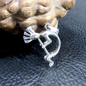 May include: A silver pendant featuring the Kokopelli figure, a symbol of fertility and music in Native American cultures. The pendant depicts the humpbacked flute player with a stylized design. The pendant is displayed against a dark background.