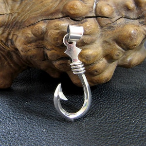 May include: A silver-toned fish hook pendant with a detailed design. The hook is curved and has a small point at the end. The pendant is hanging from a wooden surface.