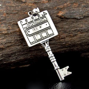 May include: A silver-colored pendant in the shape of a key. The key's head is a rectangular plate with engraved Hebrew text and symbols. The key's handle is detailed with a series of rounded shapes. The pendant is displayed on a dark wooden surface.