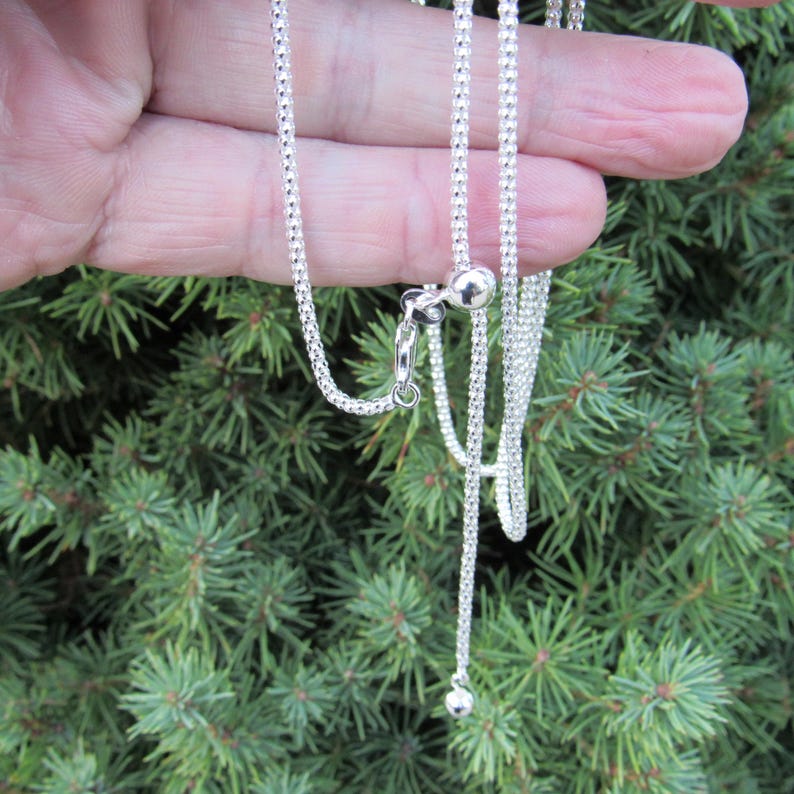 Op de afbeelding: Een zilveren ketting met een kralenontwerp en een schuifsluiting. De ketting wordt getoond op een hand en is ongeveer 61 cm lang.