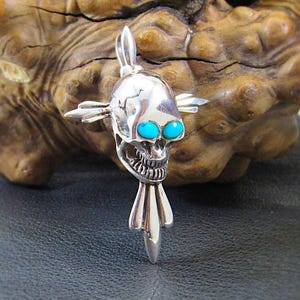 May include: A silver skull pendant with turquoise eyes and a detailed design. The skull is on a silver cross with a pointed tip.