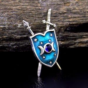 May include: A silver-toned shield-shaped brooch with a teal enamel background, adorned with stars and crescent moons. A purple gem is set in the center. A sword and spear are crossed behind the shield, creating a unique, fantasy-themed accessory.