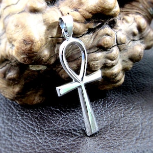 May include: A silver-colored ankh pendant, a symbol of eternal life, is displayed against a textured wooden background. The pendant features a loop at the top and a cross shape below. The metal has a polished, reflective surface.