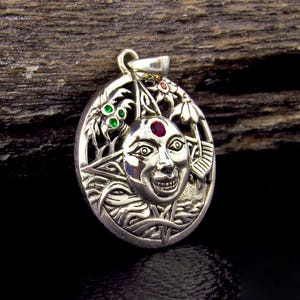 May include: Oval silver pendant featuring a smiling sun face with a red gem on its forehead. The pendant has intricate details, including floral and leaf designs, and small green gems. The pendant is suspended from a silver loop.