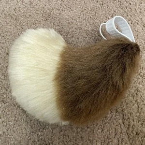 Puede incluir: Una cola de piel sintética marrón y blanca con un lazo elástico blanco para sujetar a un cinturón o cintura.