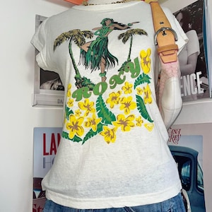 Puede incluir: Camiseta blanca con un gráfico colorido de una bailarina de hula, palmeras y flores amarillas. La palabra "Roxy" está impresa en verde. La camiseta tiene mangas cortas y cuello redondo.