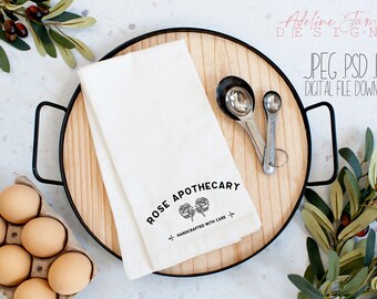 May include: A white kitchen towel with the text "Rose Apothecary" and a floral design. The text "Handcrafted with Care" is also included. The towel is on a wooden tray with measuring spoons.