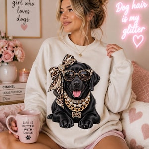 May include: A cream-colored crewneck sweatshirt featuring a graphic of a black Labrador retriever wearing leopard-print sunglasses, a matching leopard-print bow, and layered gold and pearl necklaces with a bone-shaped pendant.