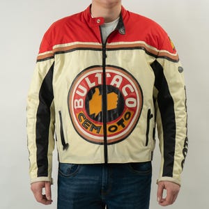 Può includere: Una giacca da motociclista bianca, rossa e nera con un grande logo "Bultaco Cem Moto" sul davanti. La giacca ha una chiusura a zip e due tasche con zip sui lati.