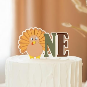 May include: A white frosted cake on a wooden cake stand, topped with a turkey and the word "ONE" in green and brown letters. A small pumpkin is to the right of the cake. The cake is likely for a first birthday celebration.